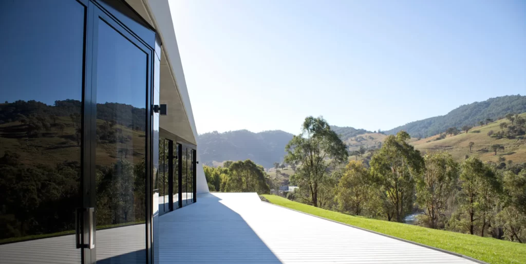 Facade of a contemporary house showing bushland reflected in the floor to ceiling glazing and a gleaming white deck
