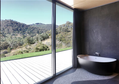 Bath tub in the dark tiled accessible bathroom. Full height windows with deck and view beyond.