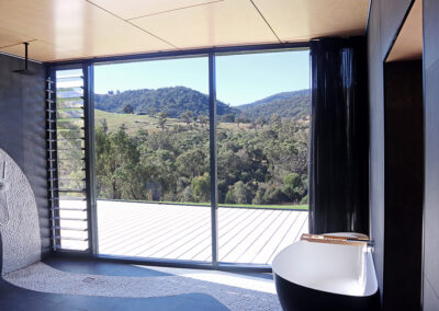 Accesible Bathroom showing mosaic tiles on the floor and a bathtub overlooking deck and rural view. Curtains are open.