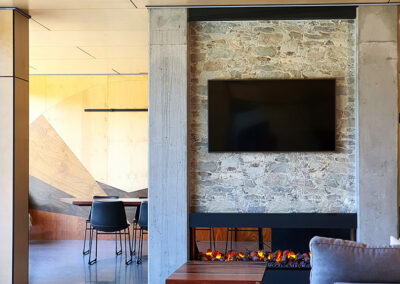Fireplace made from stone and concrete with fan hanging from ceiling. Foreground has lounge, background has dining table. All earthy tone-on-tone colours.