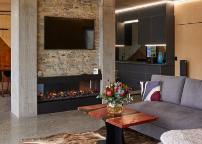 Fireplace made from stone and concrete with LED strip around the top. Foreground has lounge, background has kitchen cabinetry. All earthy tone-on-tone colours.