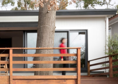 Exterior - New deck and extended facade are visible. There is a tree growing through the timber deck creating striking verticality. Our client is a blurred figure in action, walking across the new part of the deck to the new steps which connect to the garden.