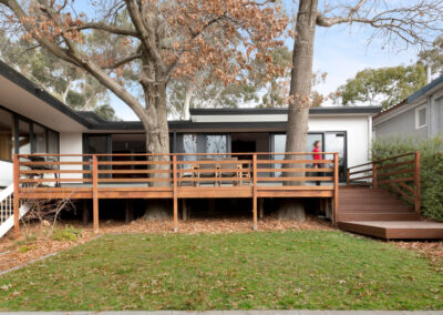 Midcentury Heritage House in Canberra: exterior shot of Neville Gruzman's midcentury modern house. New deck and extended facade are visible. There are two trees growing through the timber deck. Our client is a blurred figure in action, walking across the new part of the deck to the new steps.