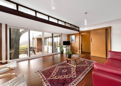 Midcentury Heritage House in Canberra: Neville Gruzman's Kennard House. Lounge room space with highlight windows and access to bedrooms via original timber sliding door. Full height windows below looking out over deck and view.