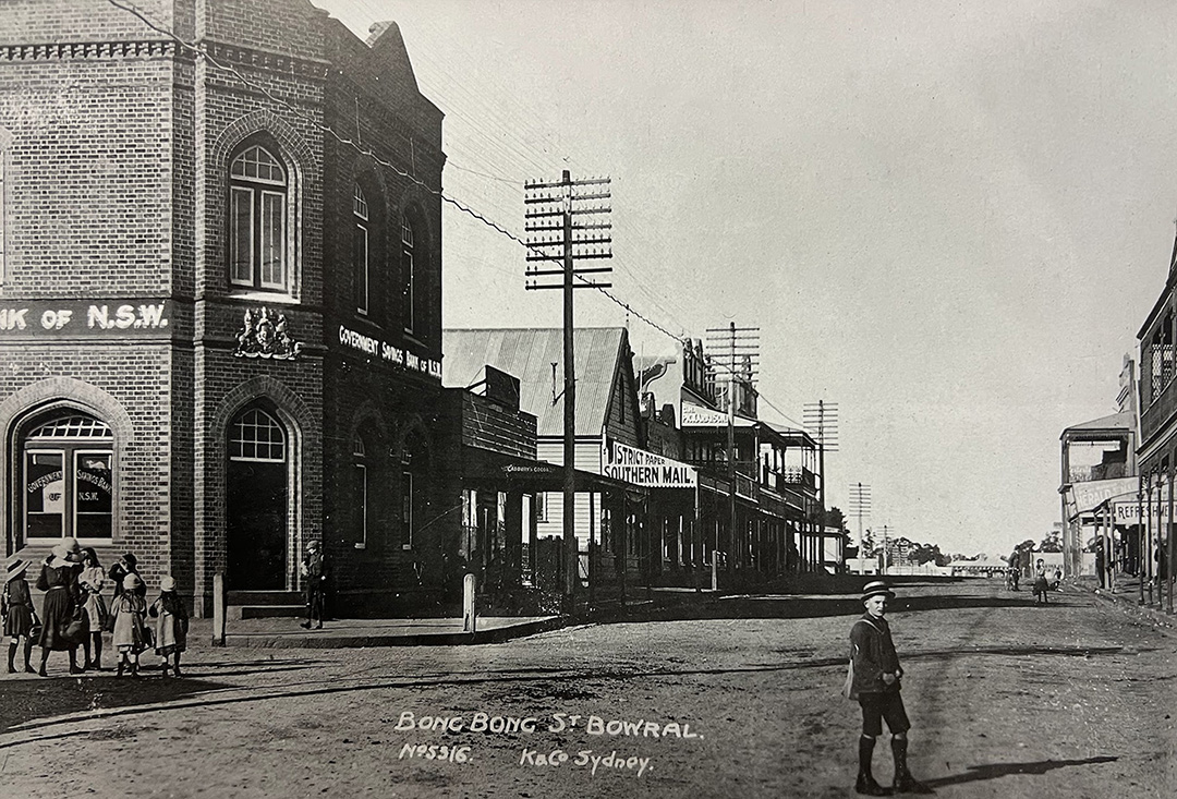 Black and white photo from 1900 showing Bong Bong St in Bowral. You can see the old bank with some smaller timber structures behind.